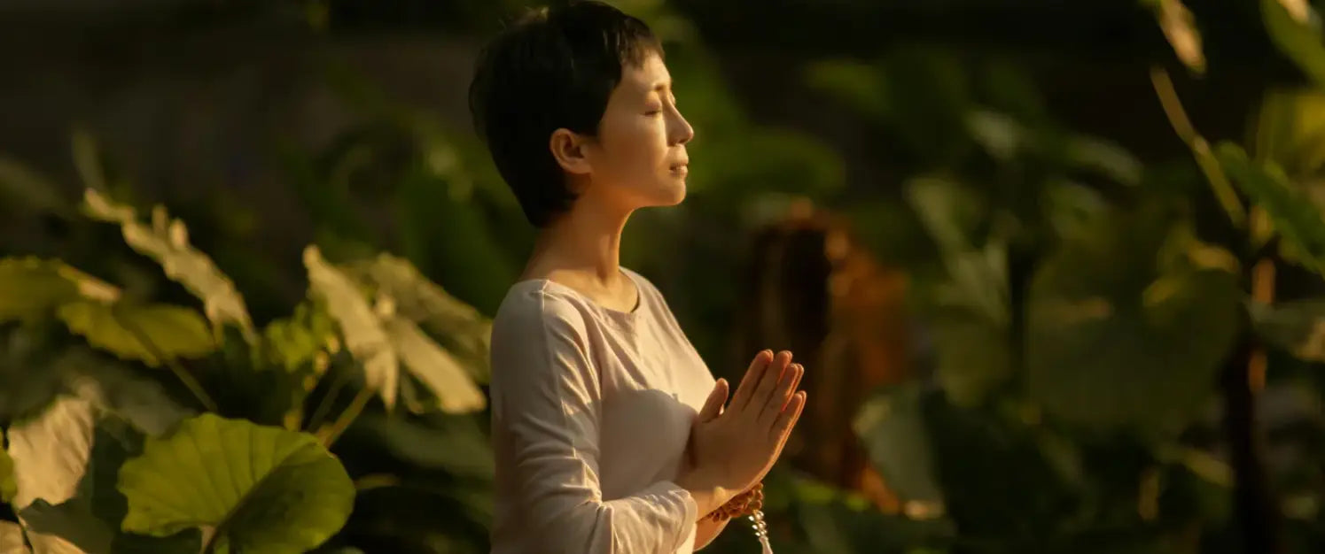 Woman meditating outdoors in a peaceful garden setting, sitting cross-legged with hands in prayer position surrounded by green plants.