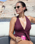 Woman sitting on the white seats of a boat, wearing a burgundy swimsuit and sunglasses, looking to the side. In the background, the blue water of a lake and rocky cliffs can be seen under the sunlight. In her hand, she holds a small pink square package that reads "Glow Beauty + Collagen Strips".