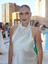 A woman wearing a white satin halter-neck dress stands outdoors by a pool with a city skyline in the background.